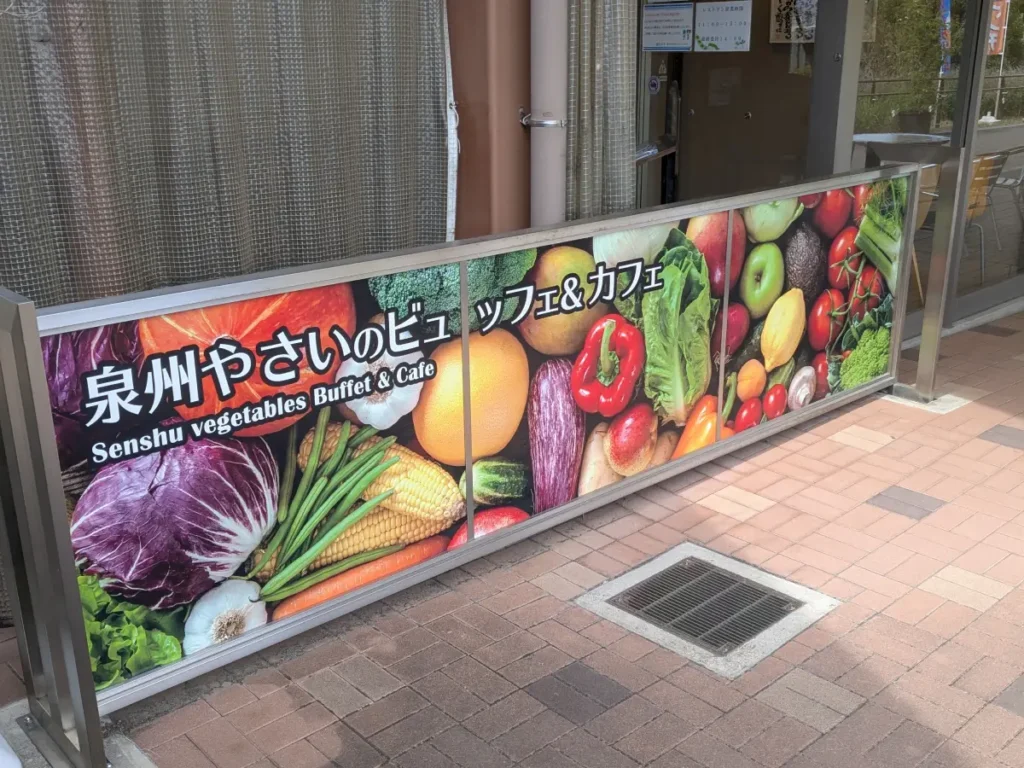 愛彩ランド入口の「泉州やさいのビュッフェ＆カフェ」看板。カラフルな野菜が描かれた目を引くデザイン